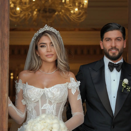 Bride in a white wedding dress and veil, holding a bouquet, standing next to a groom in a black tuxedo with a chandelier in the background.